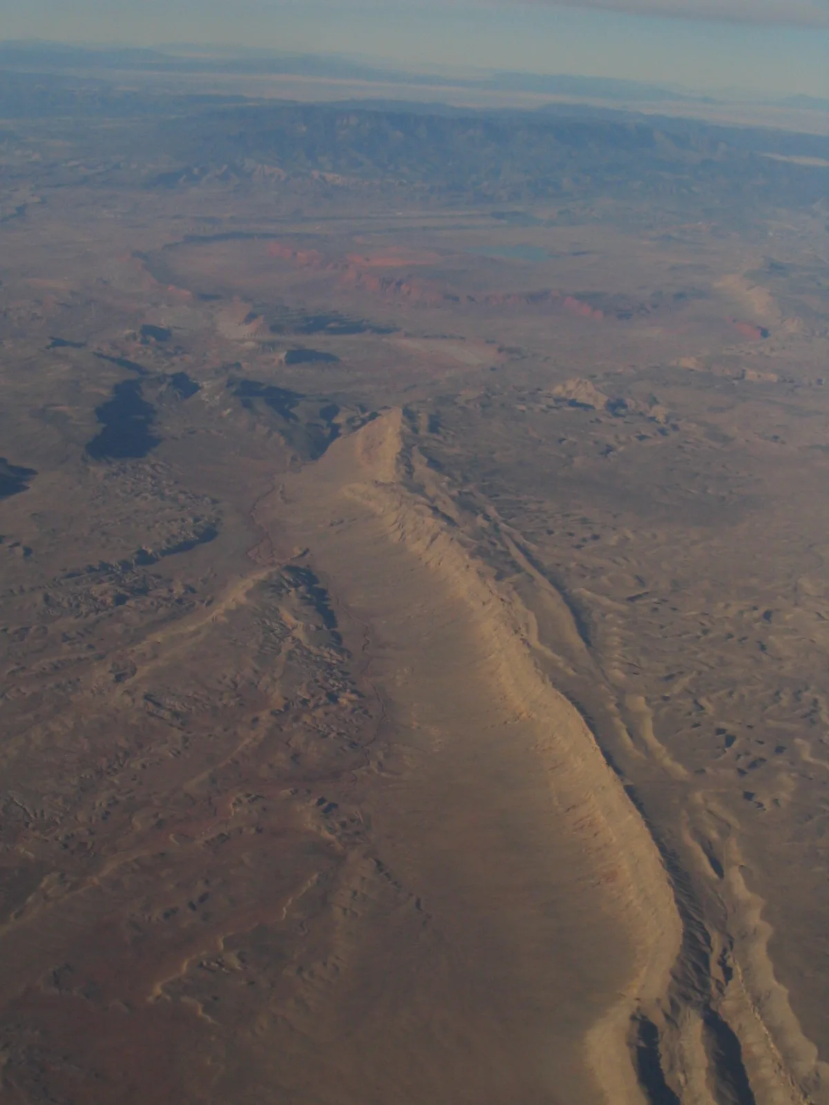 Golden-hour light washes over a long, pale sandstone ridge cutting diagonally through a vast desert basin, with layered mesas and a hazy blue mountain range in the distance. The scene feels expansive and dry, with soft shadows tracing the ridgeline and reddish cliffs faintly glowing on the horizon.