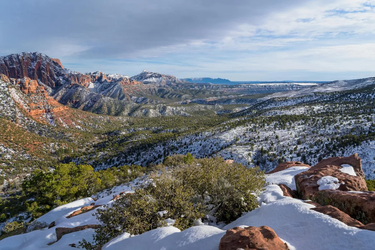 Snow-dusted red rock outcrops and scrub oak frame a sweeping overlook of the Animas Valley, with layered ridges and cliffs stretching toward the horizon under a pale blue sky. The winter light brings out the orange sandstone and green pines across the valley floor.