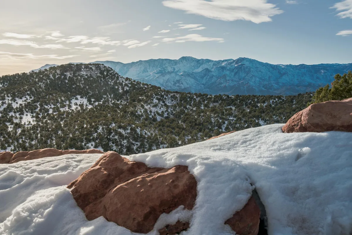 Snow-dusted red rocks fill the foreground, overlooking a dark green scrub-oak hillside and the wide Animas Valley beyond. Blue La Plata Mountains stretch across the horizon under a bright sky with thin, glowing clouds in late-day light.