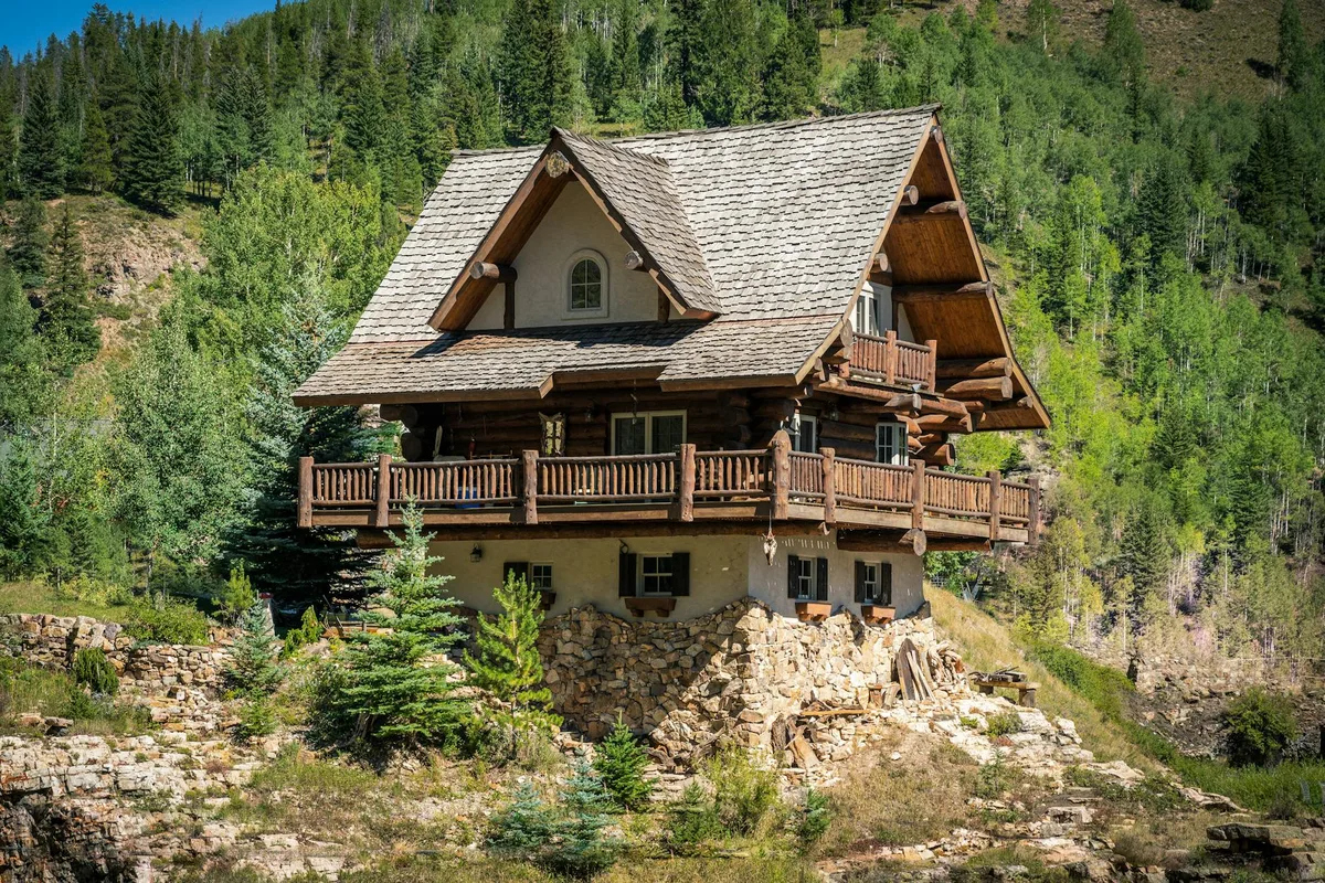 A rustic log lodge with steep shingled gables and wraparound wooden decks sits on a rocky slope in a green alpine valley. Sunlight brightens the timber details and stone foundation, with dense conifers and hillsides filling the background.
