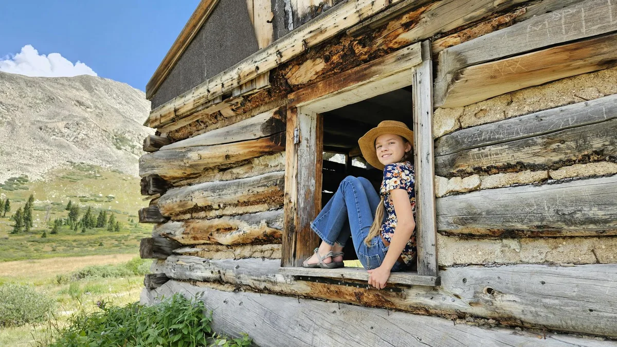A young girl in a straw hat sits in the open window of a weathered log cabin, with sunlit alpine meadow and pale rocky mountains stretching behind her. The rustic wood, bright blue sky, and green grasses give the scene a warm mountain-ranch feel.