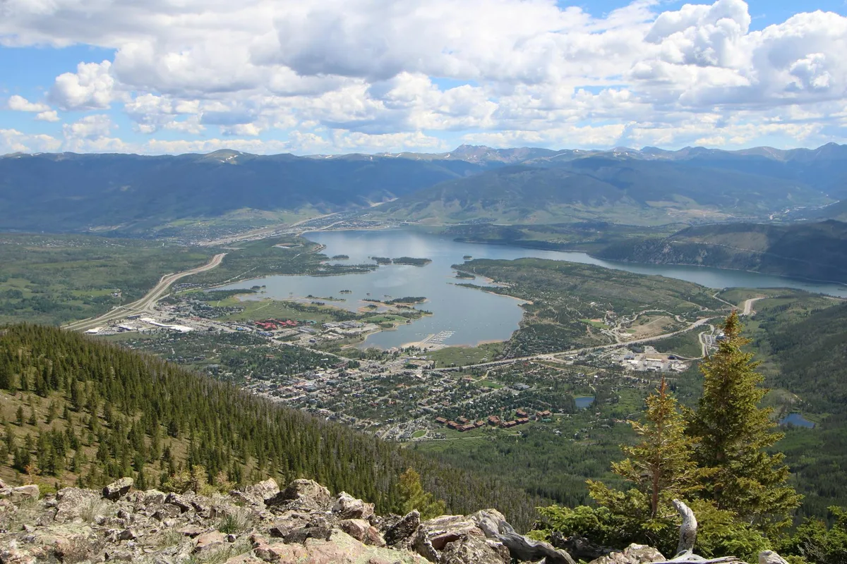 From a rocky alpine overlook, Dillon Reservoir fills the valley below, with the town spread along its shoreline and highways curving through the basin. Dark green spruce-covered slopes frame the foreground, while layered mountain ridges and scattered clouds stretch across the horizon.