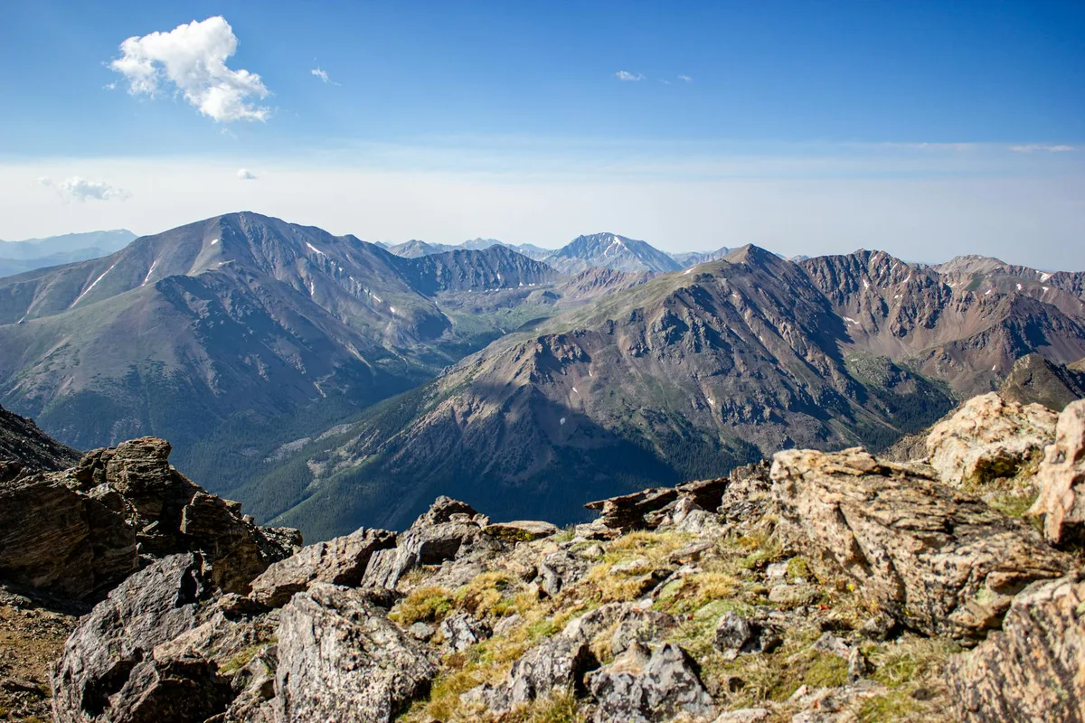 Rocky alpine tundra fills the foreground, with jagged gray boulders and tufts of grass leading to a sweeping view of rugged mountain peaks. Bright morning light and a clear blue sky with a few small clouds give the distant ridges crisp definition.