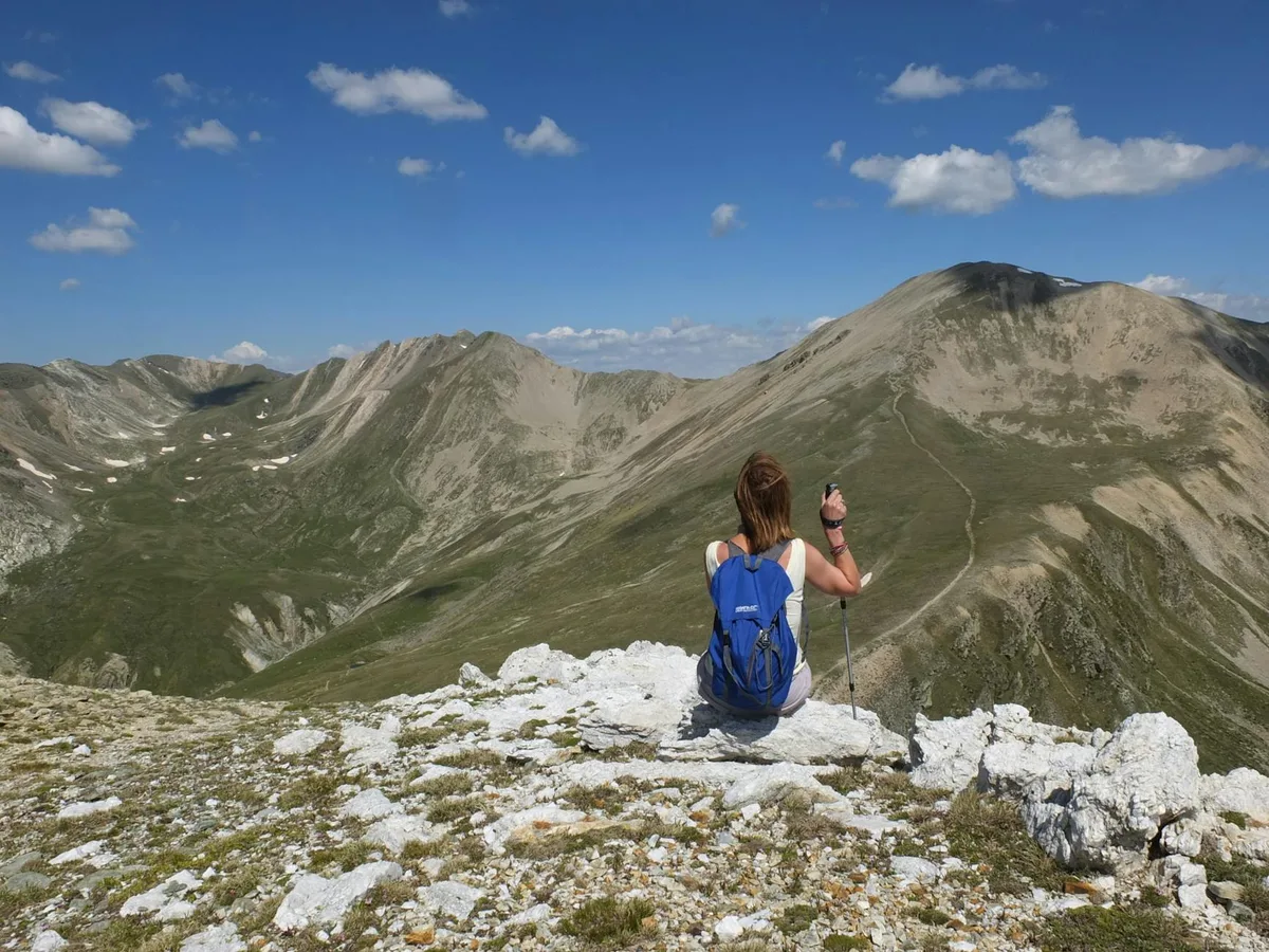 A hiker with a blue backpack sits on a rocky alpine summit, looking out over sweeping, gray-green mountain ridges under a bright blue sky with scattered clouds. The high-elevation tundra foreground and long ridge trail emphasize the wide, exposed overlook.