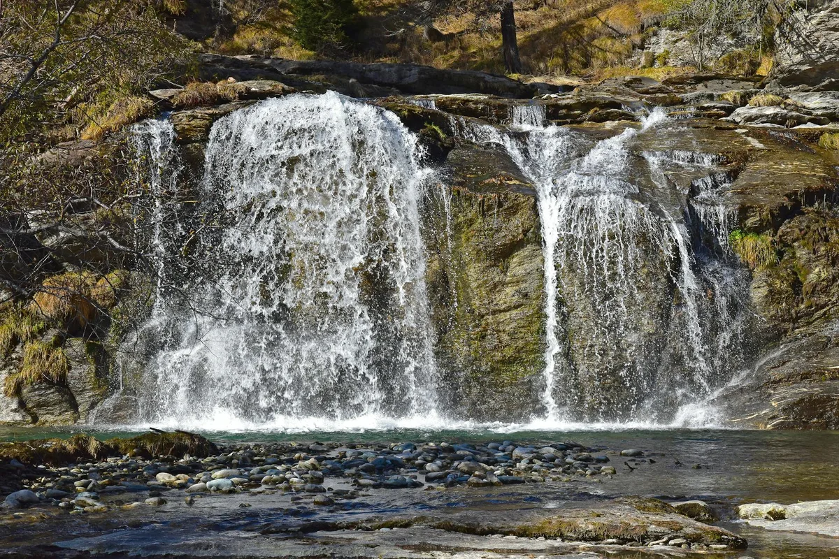A small waterfall spills over a rocky ledge into a clear pool, with white water fanning across dark stone and a pebbled streambed in the foreground. Sunlit moss and sparse alpine vegetation frame the falls, giving the scene a crisp mountain feel.