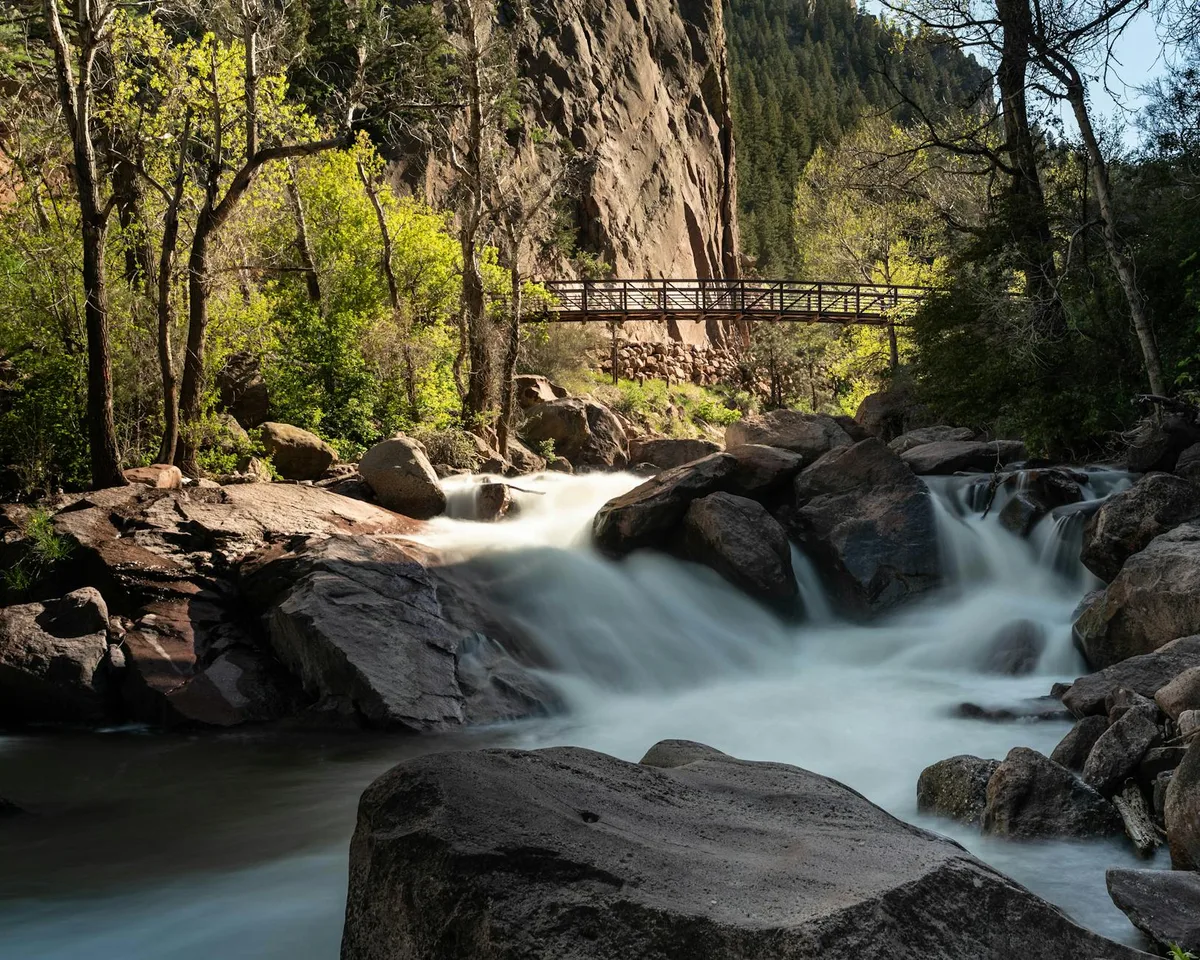 Sunlight filters through tall trees onto a rocky stream where silky white water rushes around large boulders beneath a narrow footbridge. A sheer tan cliff and dense evergreens frame the forested canyon, giving the scene a secluded, early-morning feel.