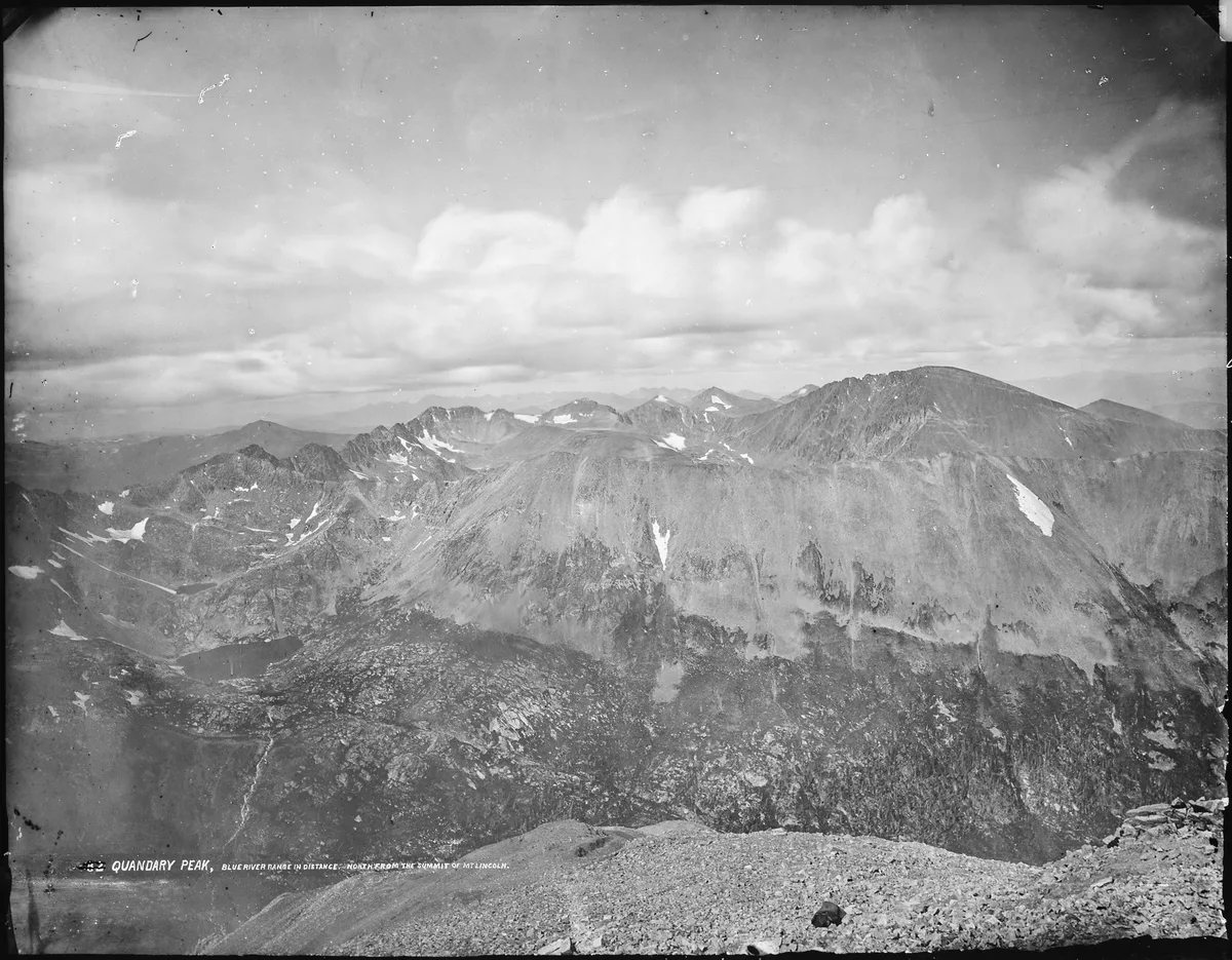 A sweeping black-and-white summit panorama shows Quandary Peak and the Blue River Range, with jagged alpine ridges, scattered snow patches, and a rocky foreground dropping away into deep mountain valleys. Thick clouds hang over the distant peaks, emphasizing the wide, above-treeline view from Mount Lincoln.