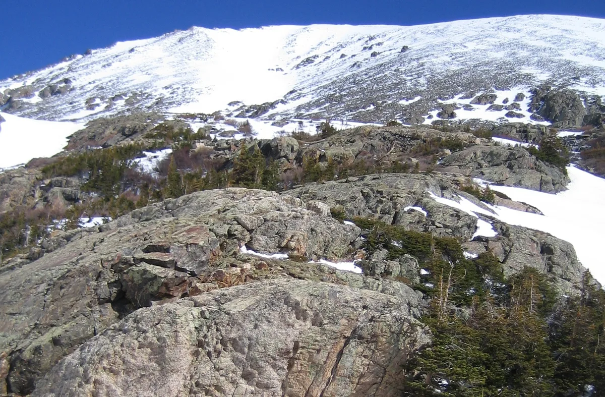 A rocky alpine ridge rises toward a broad, snow-covered summit under a deep blue sky, with scattered evergreens and patches of lingering snow across the foreground. The morning light highlights the textured granite and the steep, open above-treeline terrain.