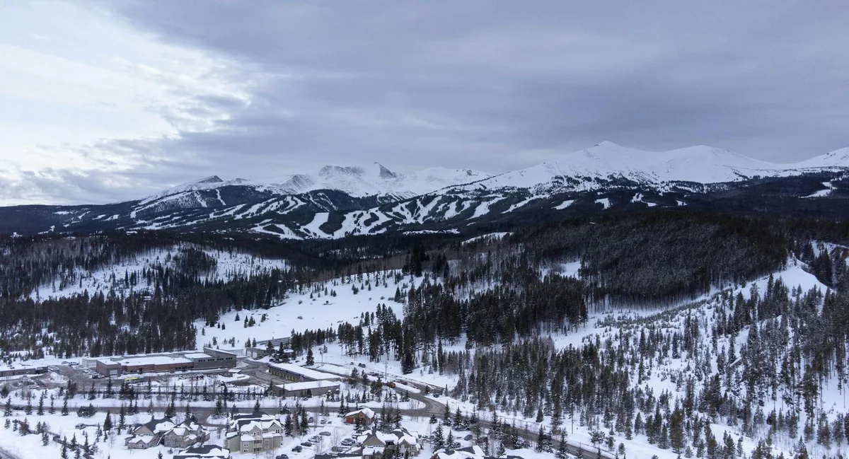 Snow-covered Breckenridge spreads across the foreground with dark pine forests and winding roads below. In the distance, the broad, white slopes of Quandary Peak and nearby ski runs stretch under a cloudy morning sky.