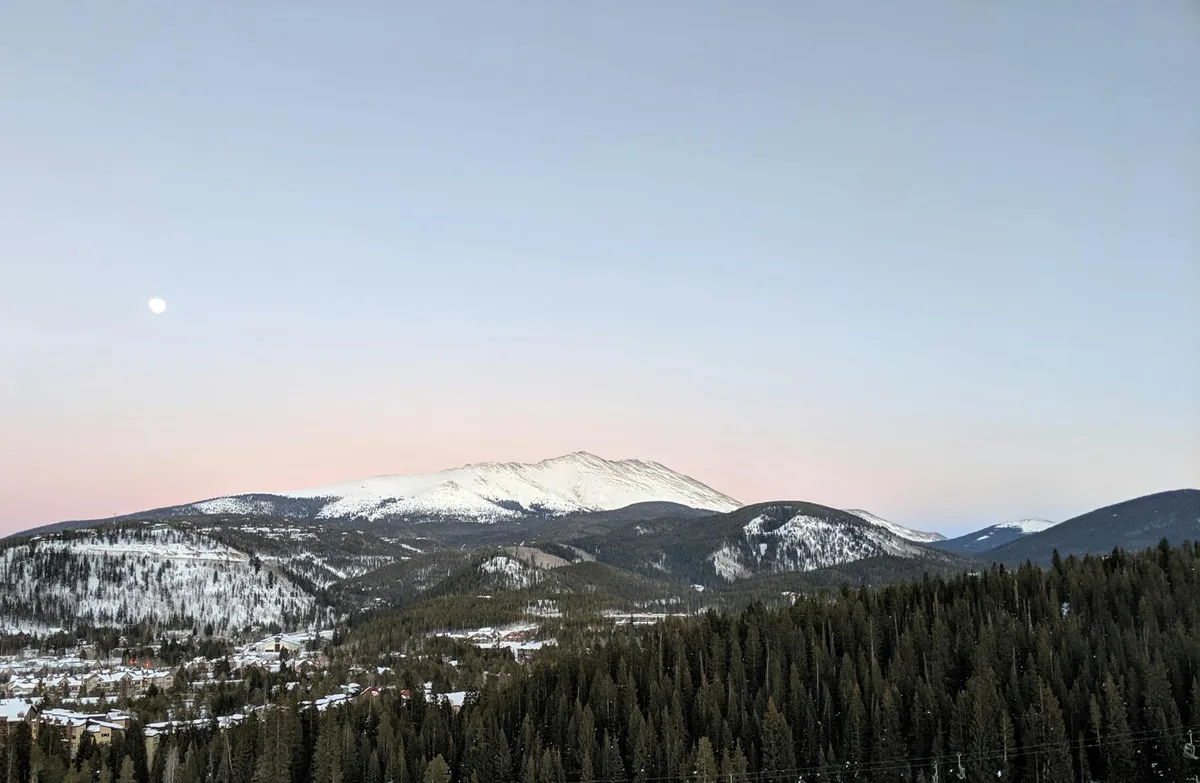 Soft morning light washes over a snow-covered Quandary Peak, with dark pine-covered hills and Breckenridge tucked in the valley below. A pale moon hangs in the clear pastel sky above the broad alpine ridge.
