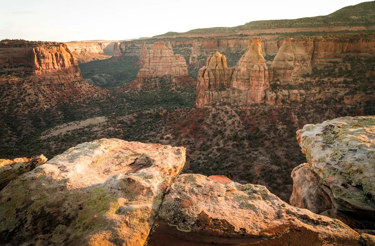 Golden light washes over a vast desert canyon of red-orange badlands and layered cliffs, with tall sculpted spires rising from the valley below. Sunlit sandstone ledges fill the foreground, overlooking the shadowed ravine and scrubby hills beyond.