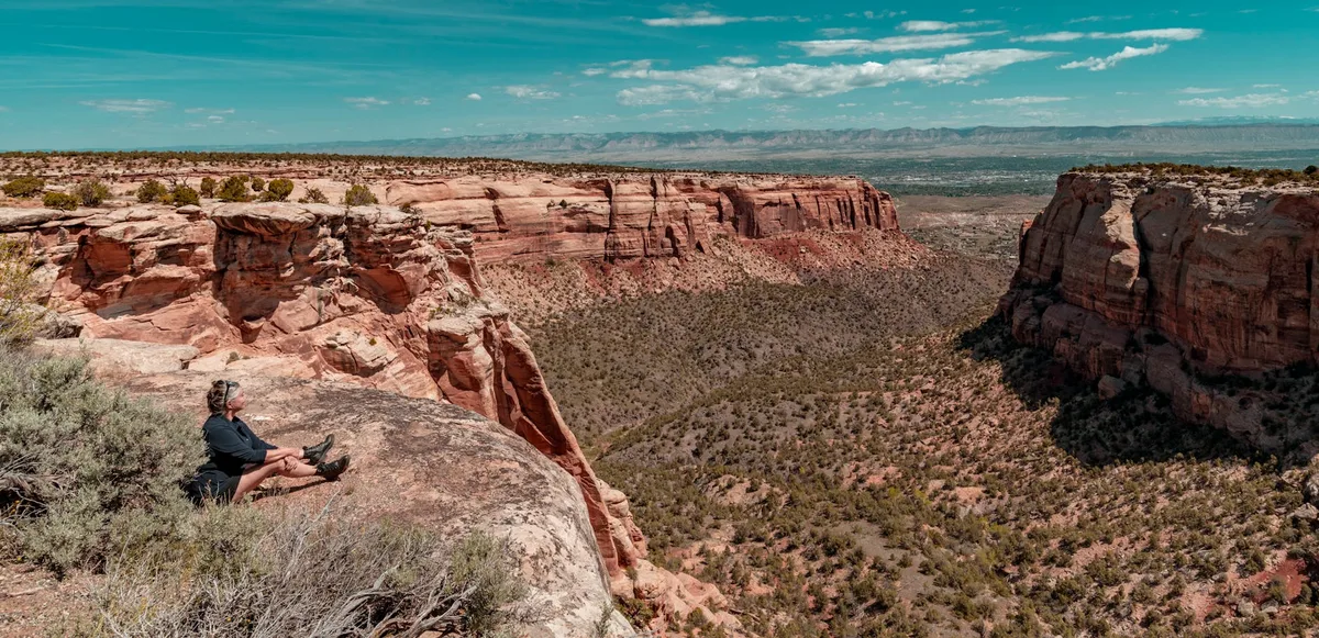 A hiker sits on a sunlit sandstone ledge above a vast red-rock canyon, with steep eroded cliffs, a juniper-speckled valley, and distant mountains stretching under a bright blue sky. The wide view emphasizes the layered badlands and rugged desert textures in the golden light.