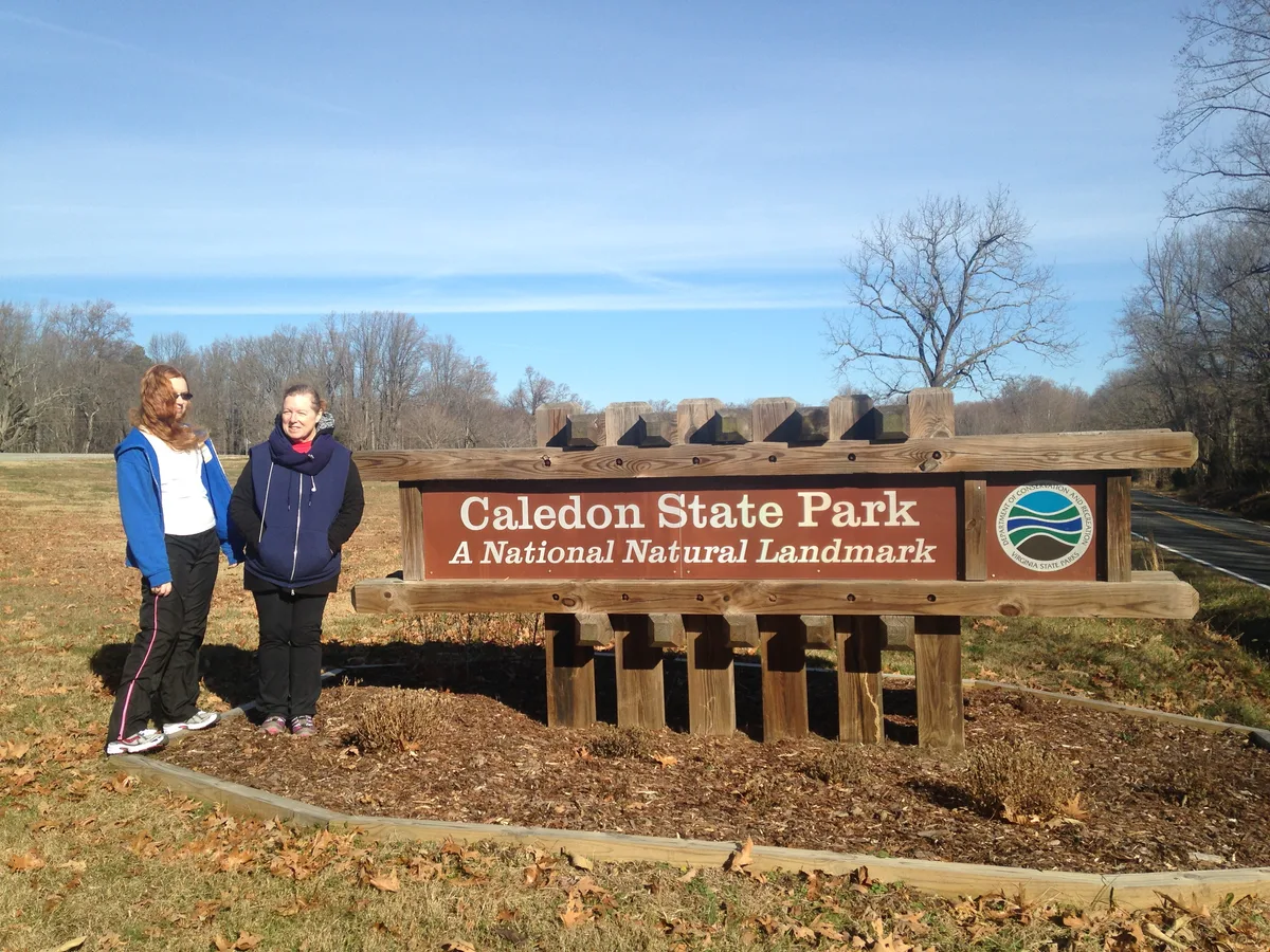 Two women stand beside a large wooden Caledon State Park sign on a sunny winter day, with leafless trees and brown grass in the background. The scene is framed by a paved road and bare landscaping, giving it a quiet off-season feel.