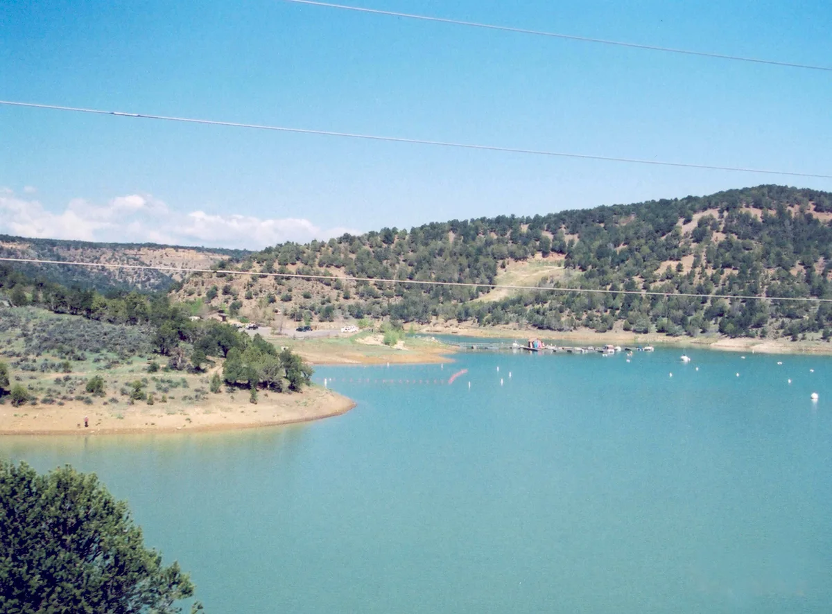 Calm turquoise reservoir water curves around a sandy shoreline and low, tree-covered hills under a clear blue sky. Small boats and white buoys dot the surface, with power lines crossing the upper part of the frame.