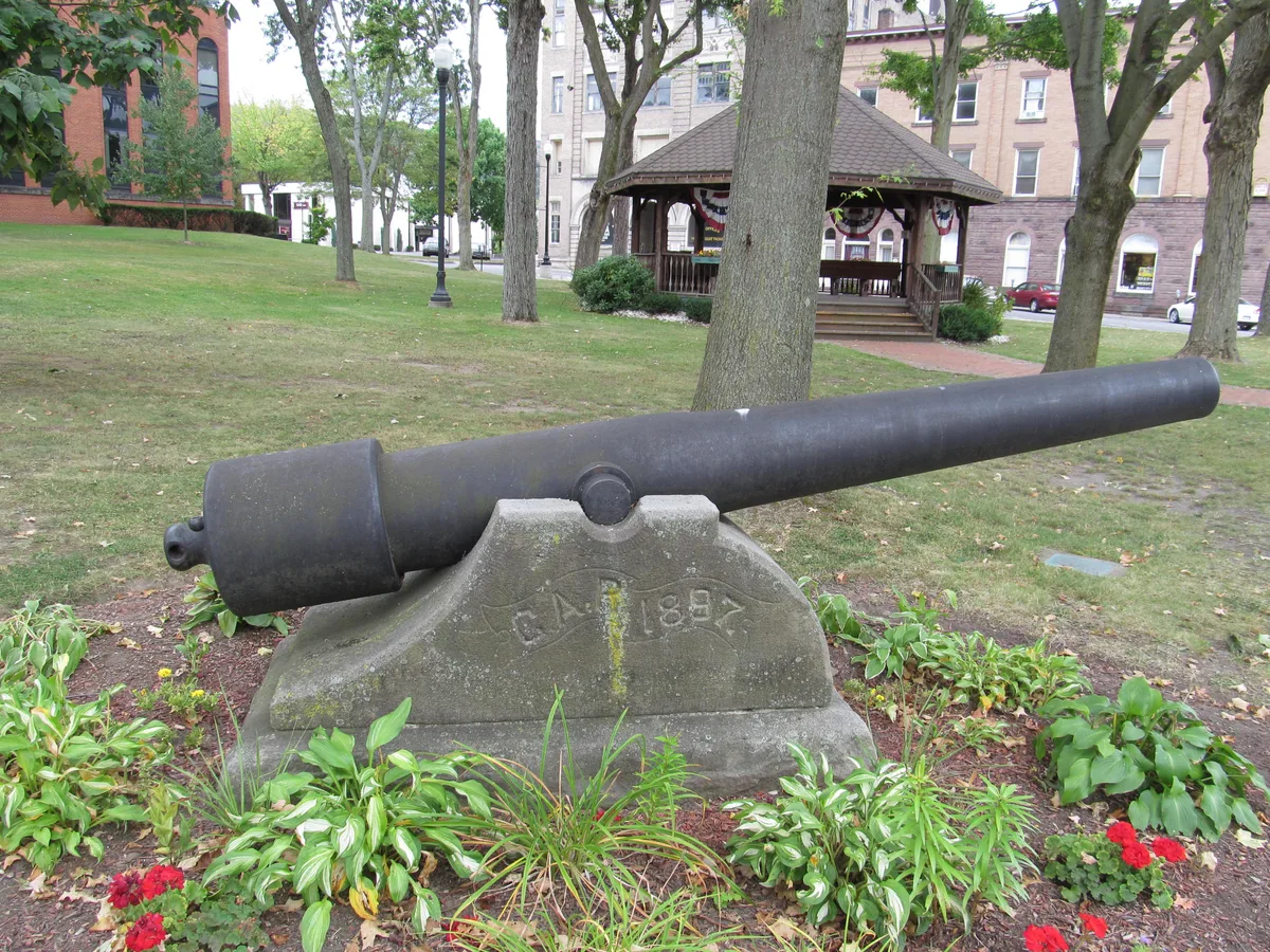 A weathered black cannon sits on a stone base in a small landscaped park, surrounded by low green plants and red flowers. Behind it, a wooden gazebo and brick buildings line the grassy square beneath mature trees.