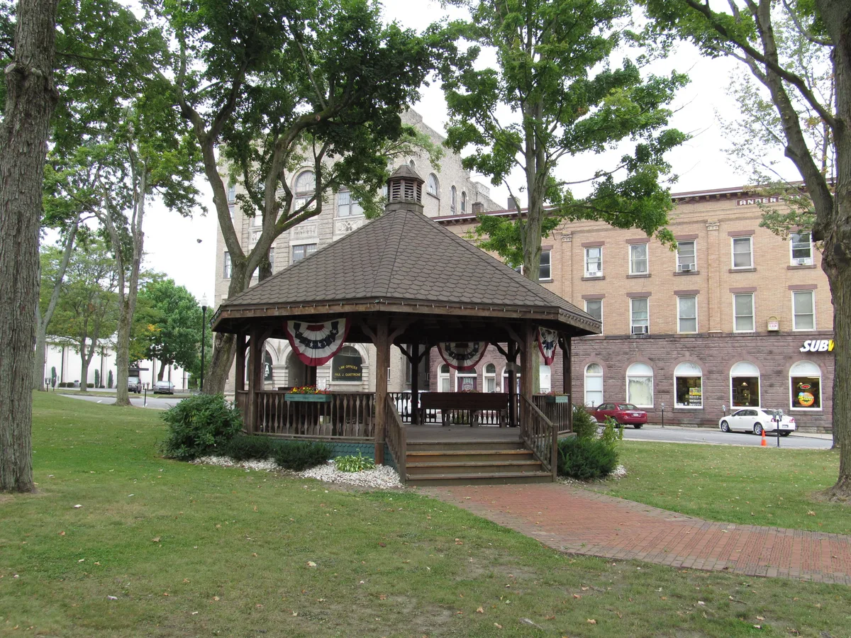 A small wooden gazebo with patriotic bunting sits in a shady grassy park, framed by tall trees and a brick walkway. Historic brick buildings with arched windows and storefronts line the background under soft daylight.