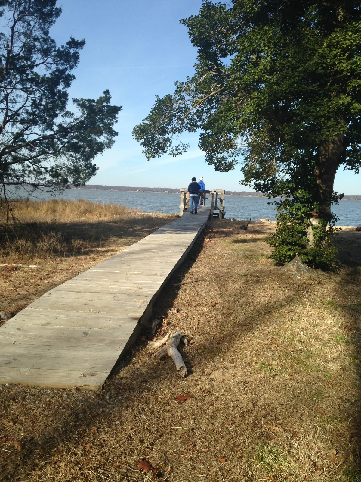 A narrow wooden boardwalk leads through dry grass and trees to the calm reservoir, where two people stand at the end looking out over the water. Clear blue sky and warm late-day light give the scene a quiet, open feel.