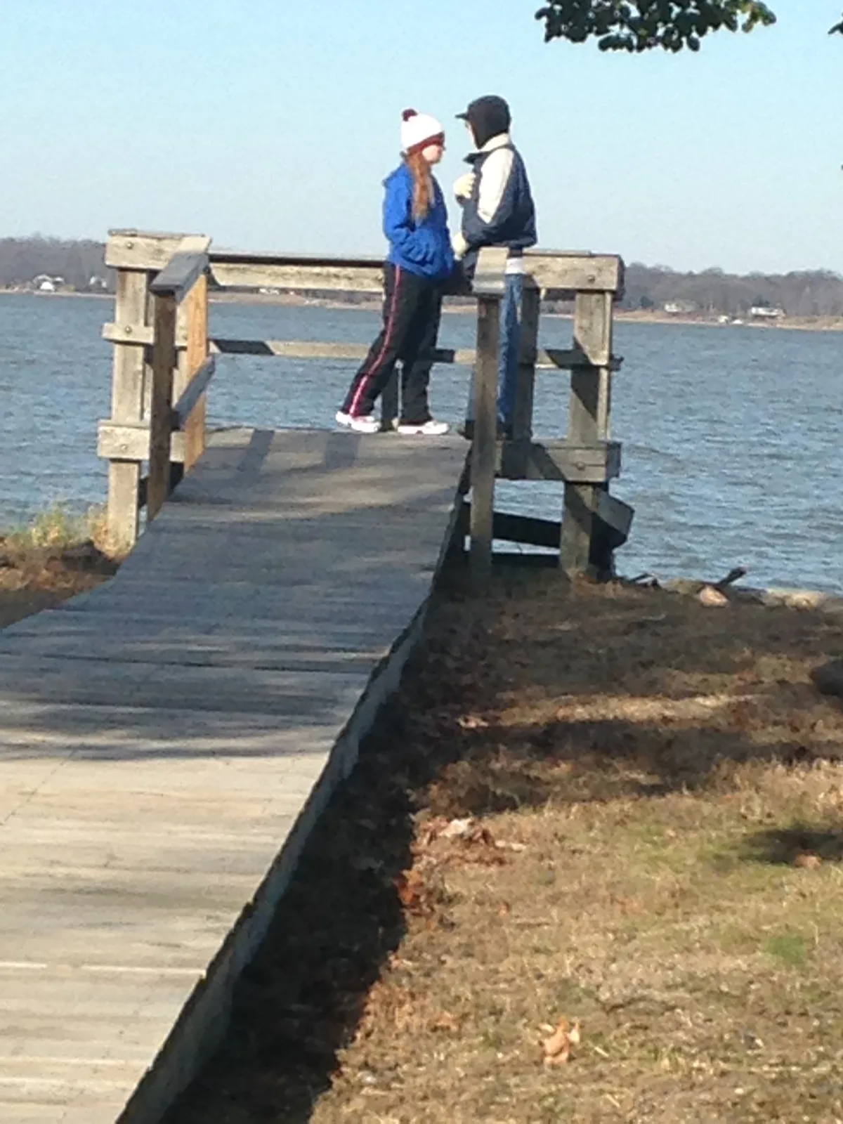 Two bundled-up people stand chatting on a wooden dock that extends into calm blue water, with a low shoreline and bare trees in the distance. Bright winter light and long shadows give the scene a crisp, open feel.