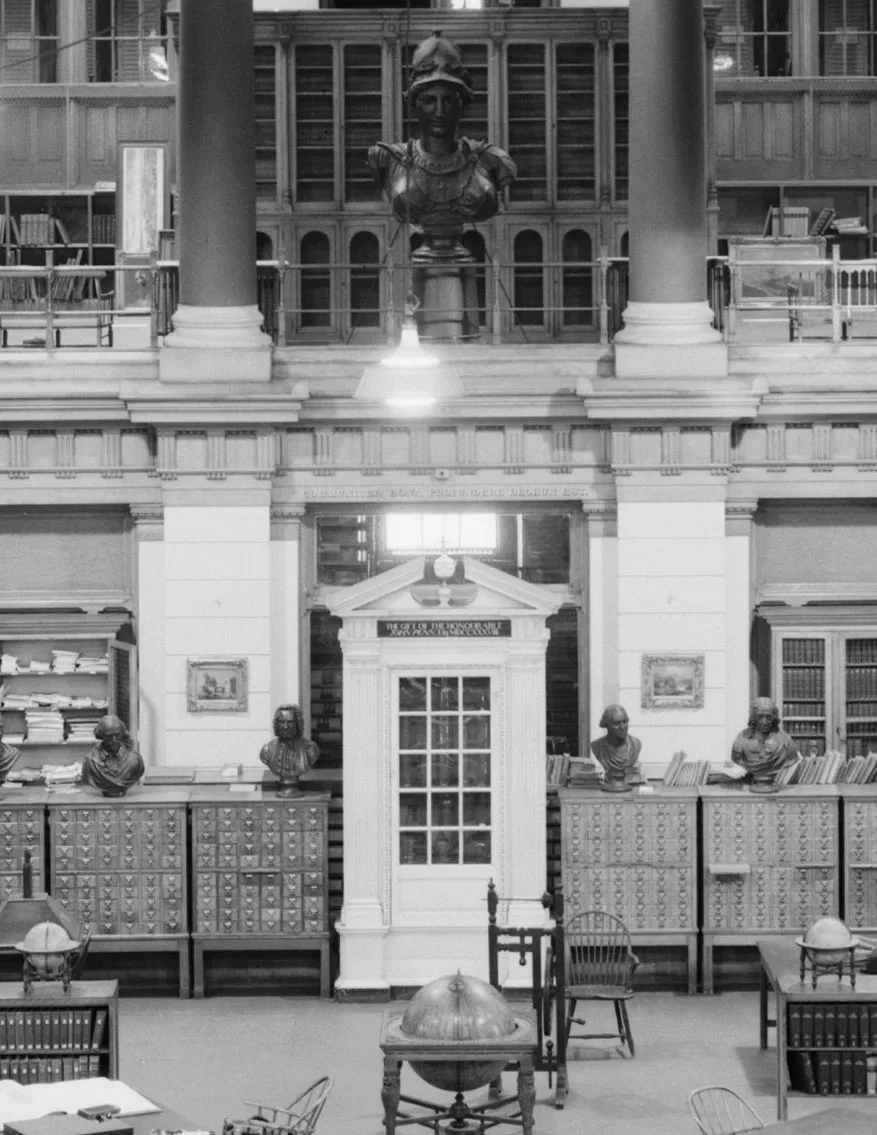 An ornate historic library interior with tall columns, a central white door, and rows of card catalog cabinets topped with busts. Shelves of books, a globe, and a large classical bust mounted above the room create a formal, old-world atmosphere.
