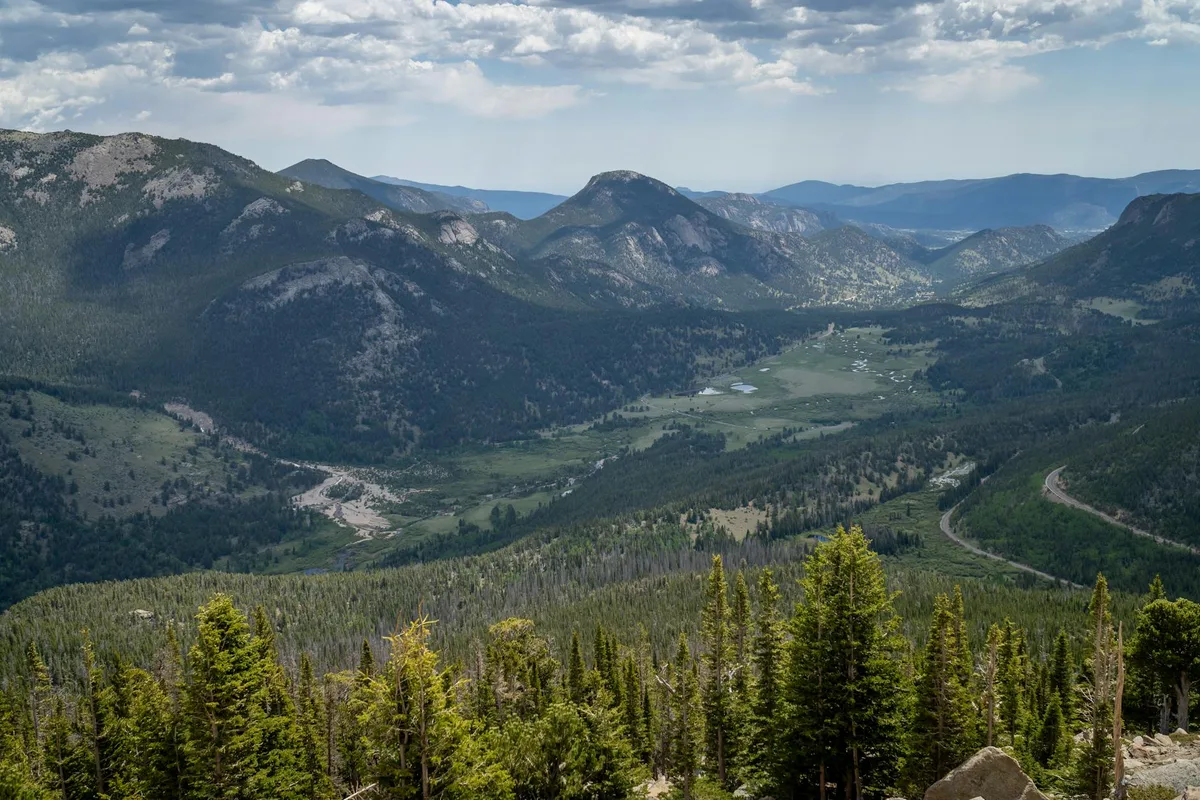 Sweeping mountain panorama with dark, rocky peaks rising above a broad green valley, viewed from a high overlook under a partly cloudy sky. Evergreen trees and a few boulders frame the foreground, with a winding road cutting across the right side of the scene.