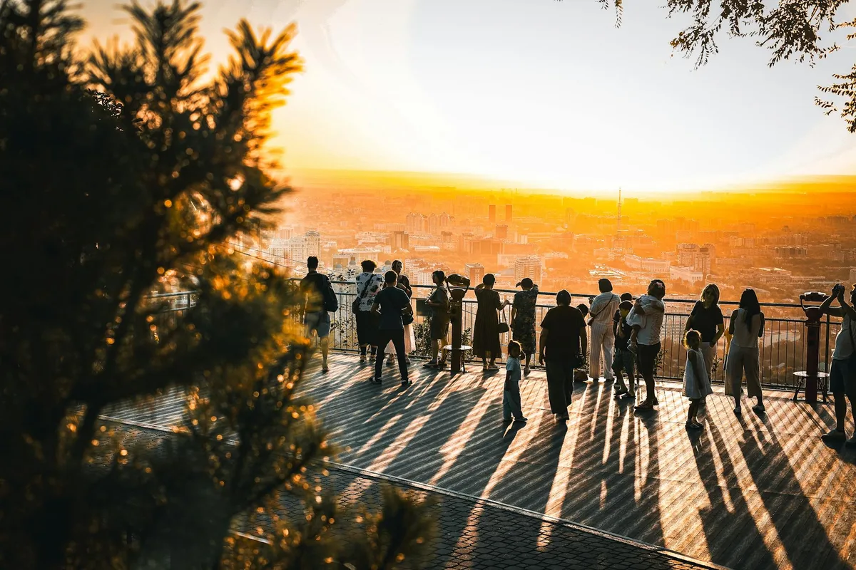 Visitors stand along a railing at a mountain overlook in warm golden-hour light, with long shadows stretching across the wooden deck. A hazy city skyline fills the distance beyond dark pine branches framing the left side of the view.