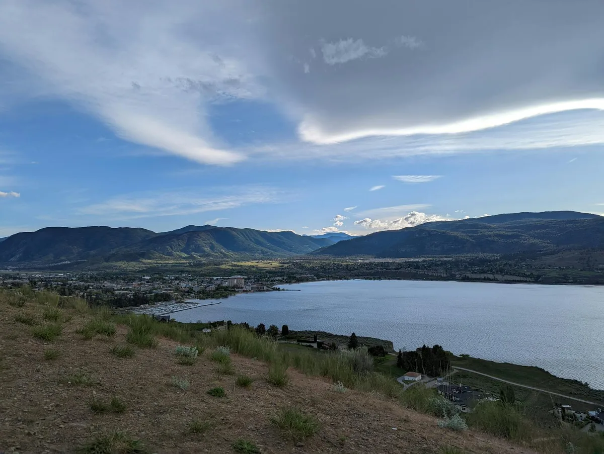 From a grassy, rocky overlook, Dillon Reservoir stretches across the right side of the frame, with the town below and dark mountain ridges lining the horizon. Soft evening light and wispy, layered clouds give the wide panoramic view a calm, open feel.