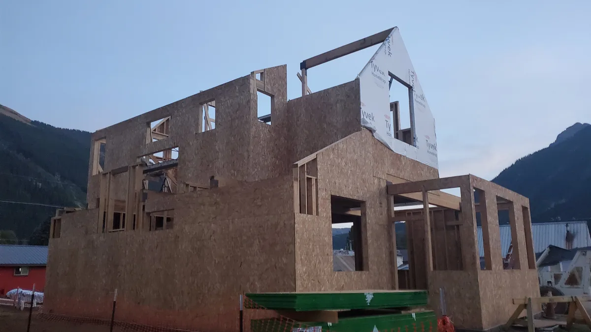 An unfinished two-story wooden building shell rises in Silverton, with exposed framing, OSB sheathing, and a white Tyvek-wrapped upper gable under a pale blue evening sky. Dark mountain slopes frame the construction site, with a few low rooftops and a small red building peeking in from the sides.