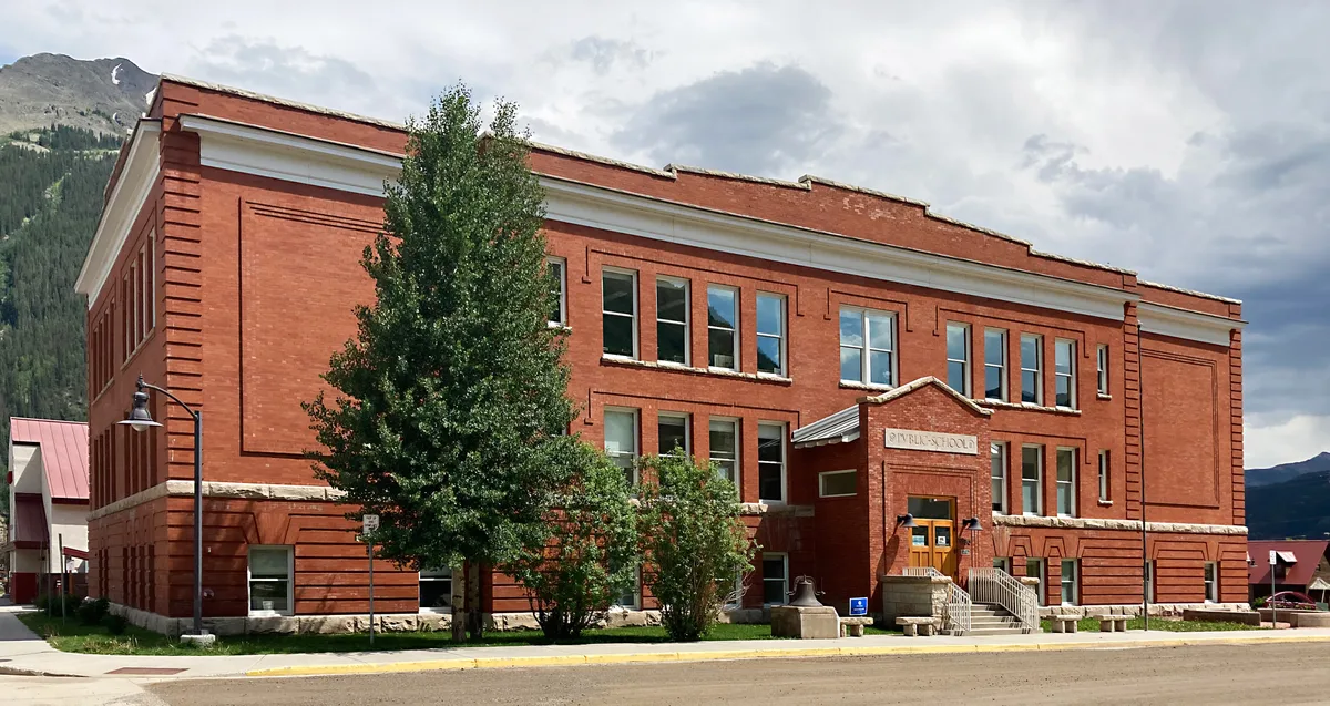 A large red-brick historic school building fills the frame, with a central entrance labeled “Public School,” white trim, and rows of tall windows. A leafy tree softens the front facade, while gray clouds and mountain slopes rise behind it in the high basin light.
