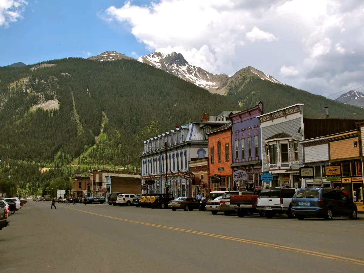 Colorful Victorian storefronts line Silverton’s main street, with parked cars and a few pedestrians in the foreground. Snow-streaked San Juan peaks rise dramatically behind the historic downtown under a bright, partly cloudy sky.