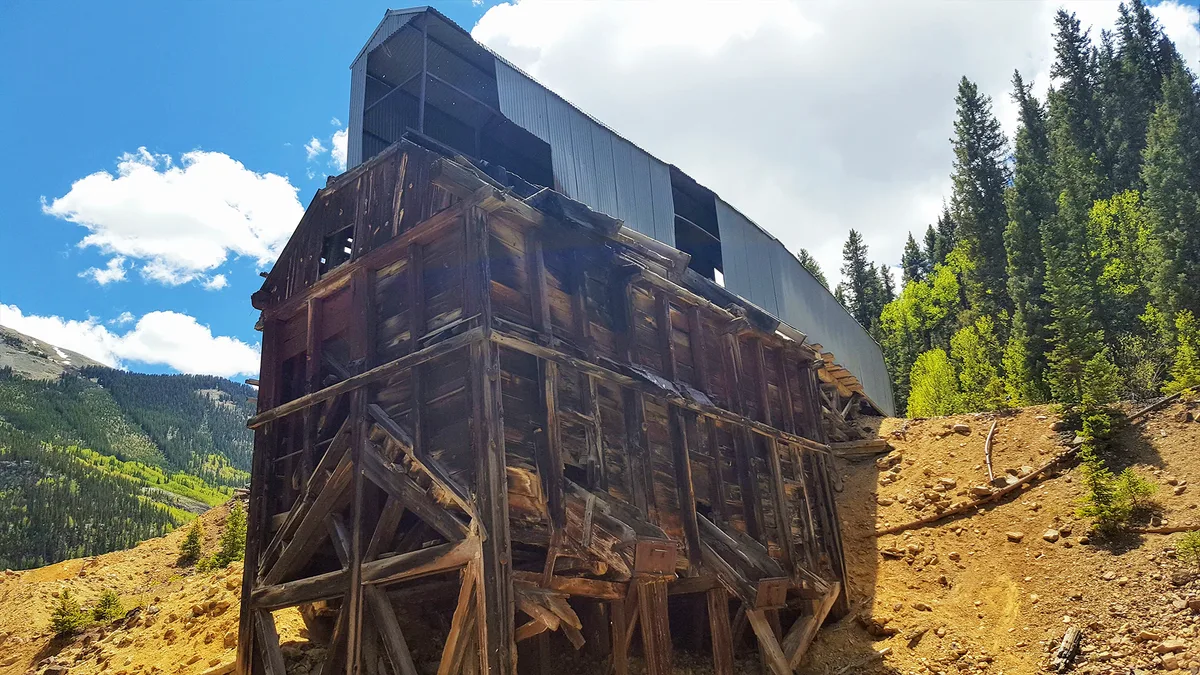 A weathered, abandoned wooden mine structure stands on a steep dirt slope, its dark, leaning timbers backed by a modern corrugated metal building. Bright midday light hits the rocky hillside, with dense green evergreens and a partly cloudy blue sky filling the mountain backdrop.