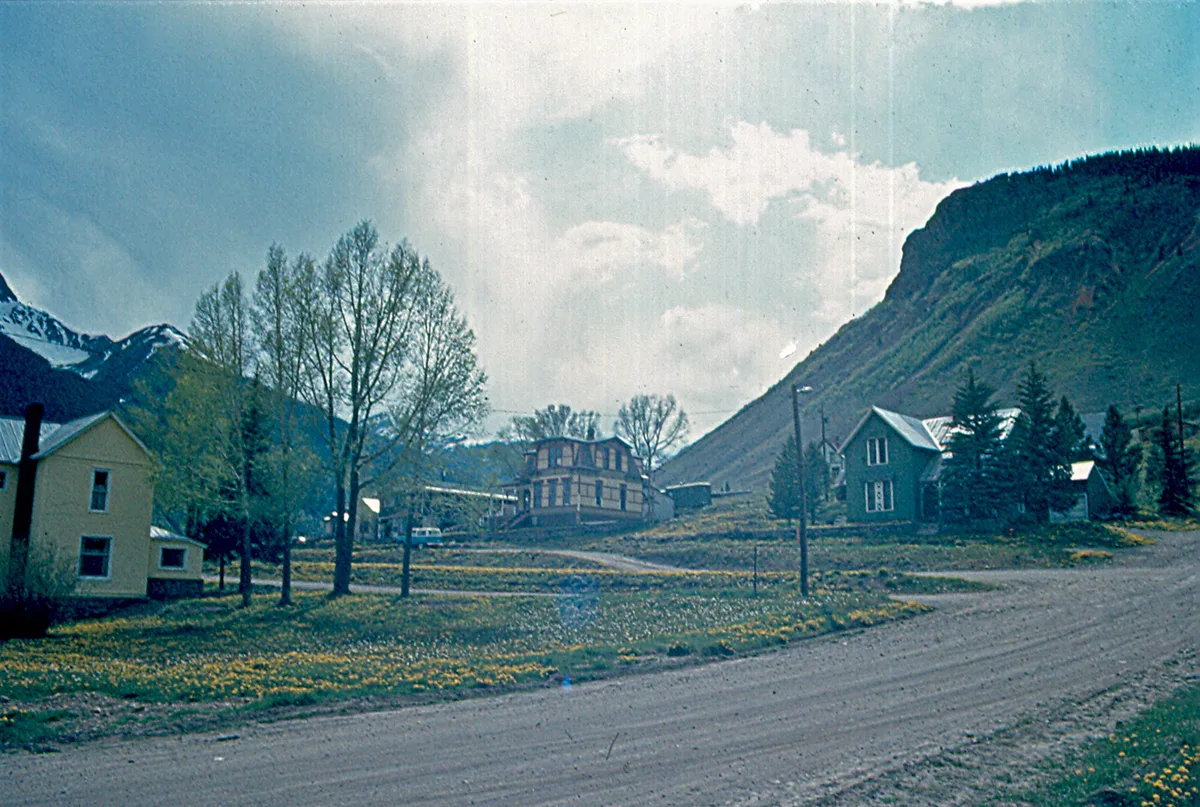 A quiet Silverton street curves through a small cluster of historic Victorian-era houses and storefronts, with bare trees and patches of spring grass in the foreground. Steep San Juan Mountain slopes rise behind the town under a bright, hazy sky.