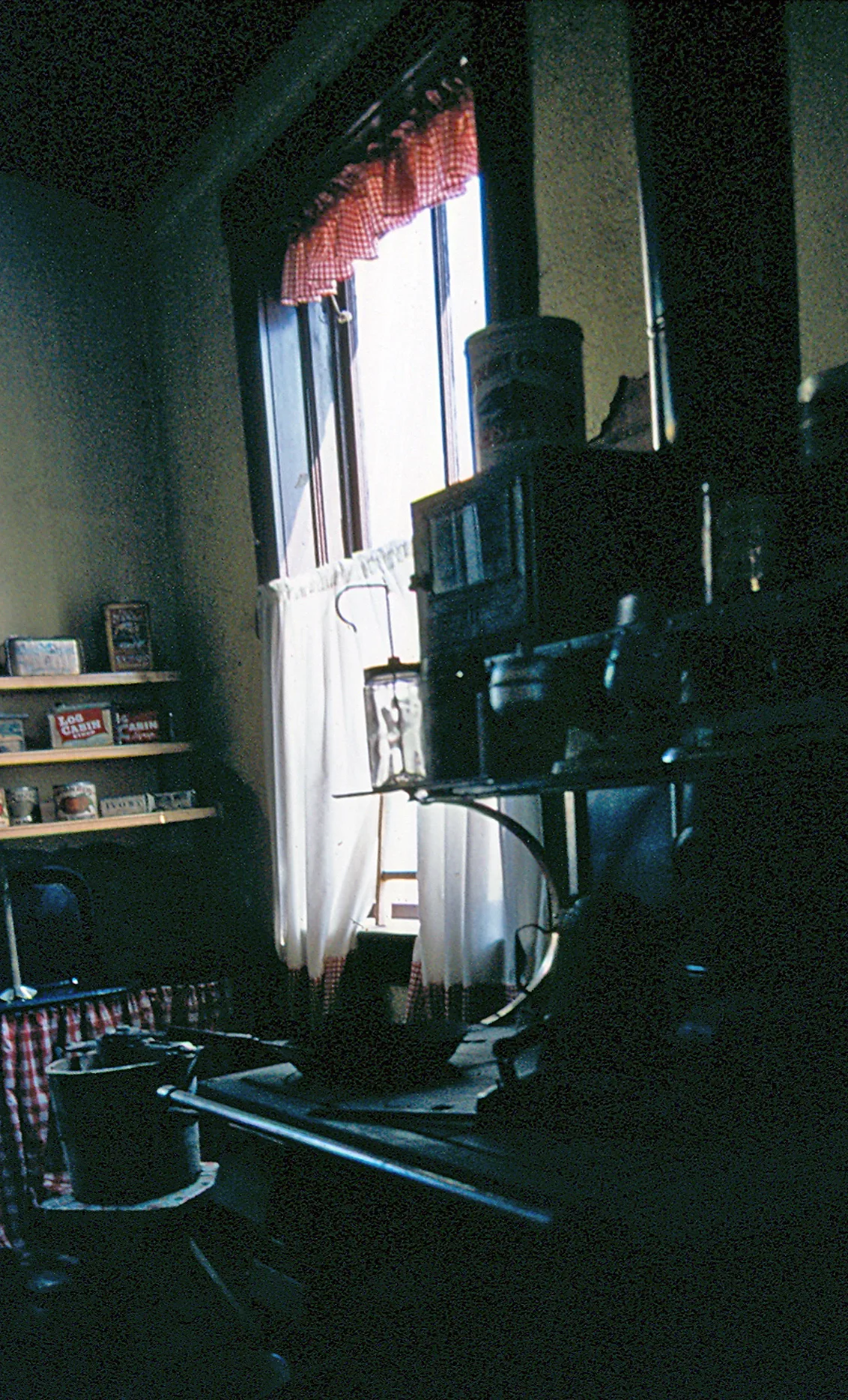 A dim, old-fashioned kitchen or pantry is lit by bright daylight through a tall window with white curtains and a red gingham valance. Shelves of canned goods line the left wall, while a dark stove and cookware fill the foreground in a cramped, cluttered interior.