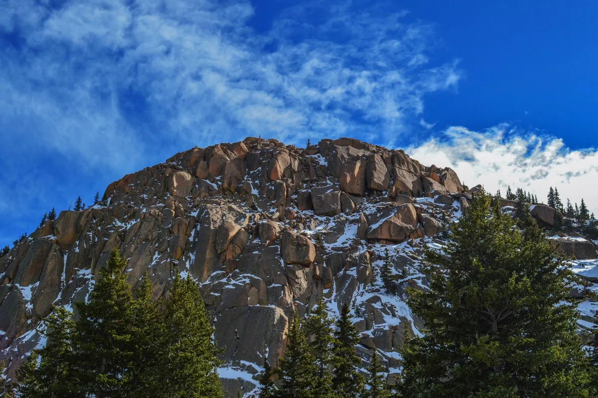 A rugged granite spire rises above dark evergreen trees, with streaks of snow clinging to the cliff face under a bright blue sky. Wispy clouds sweep across the top of the frame, emphasizing the steep, alpine landscape.