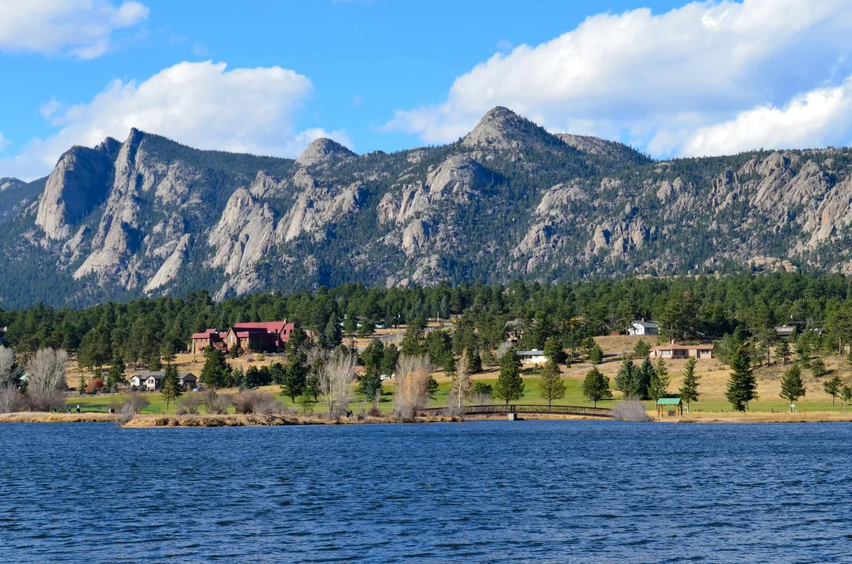A wide view across a dark blue alpine lake toward jagged granite spires and pine-covered slopes, with scattered cabins and a small shoreline bridge in the foreground. Bright morning light and puffy clouds sharpen the rocky ridge line above the water.