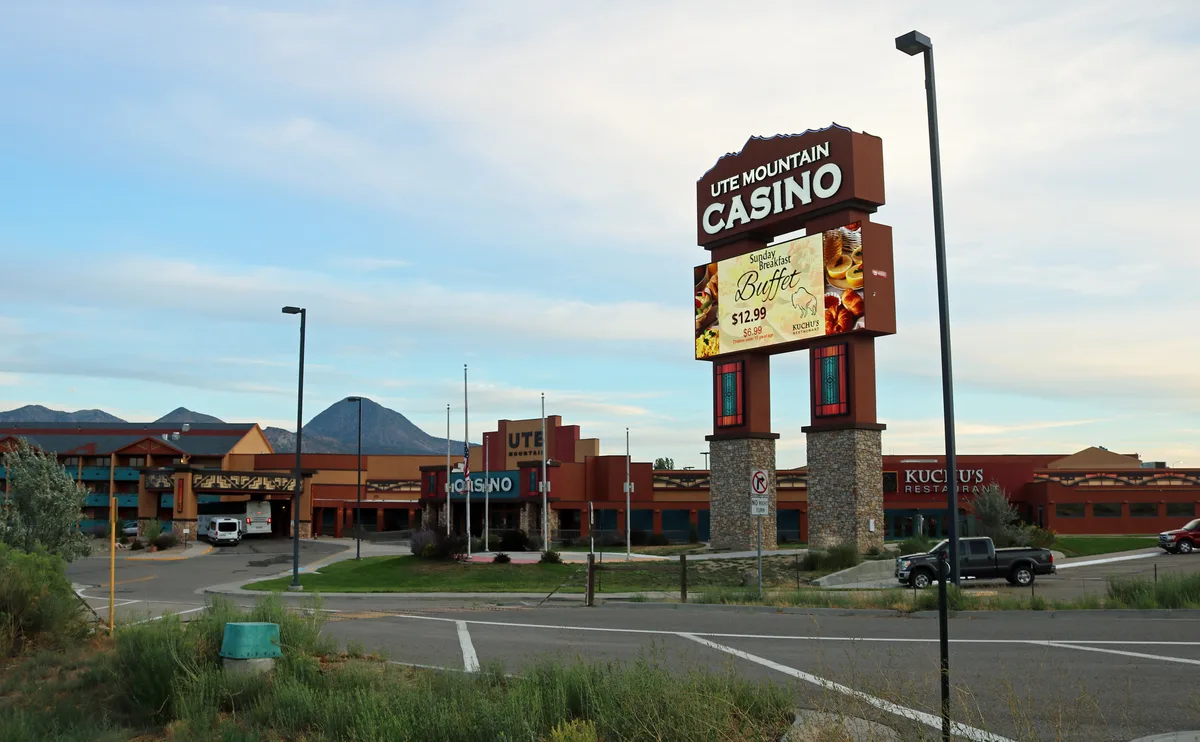 Ute Mountain Casino sits in the foreground with a tall roadside sign, parking lot, and low adobe-style buildings, while Sleeping Ute Mountain rises in blue-gray silhouette on the horizon. Soft evening light and a pale, cloud-streaked sky give the wide desert scene a calm, open feel.