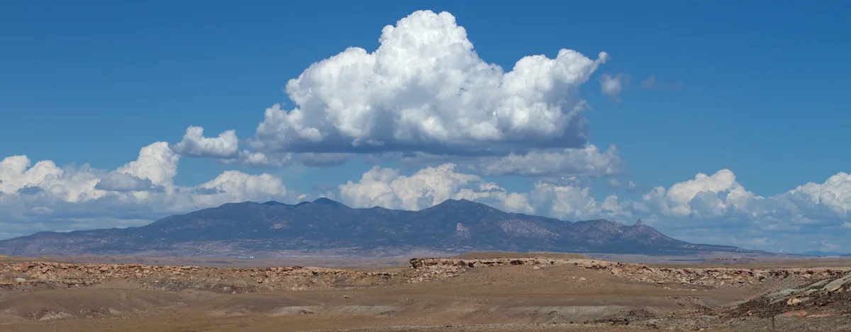 Sleeping Ute Mountain stretches low across the horizon under a bright blue sky, with a huge billowing white cloud centered above its dark silhouette. The foreground is a dry, tan desert plain with rocky ridges, creating a wide panoramic view.