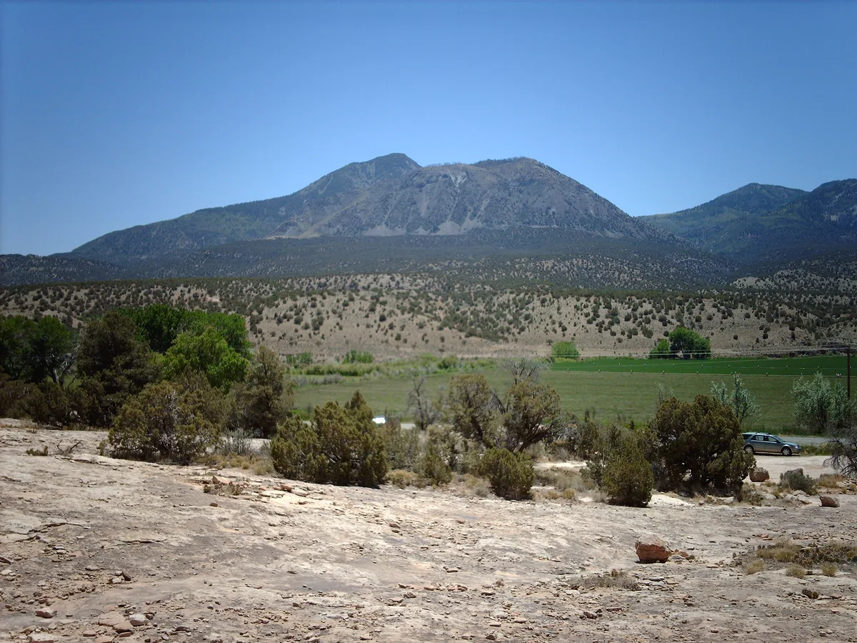 Sleeping Ute Mountain rises in a broad, rugged silhouette under a clear blue sky, with tan desert scrub and a green valley stretching across the foreground. The wide landscape view emphasizes the mountain’s layered slopes and the open, arid terrain around Towaoc.