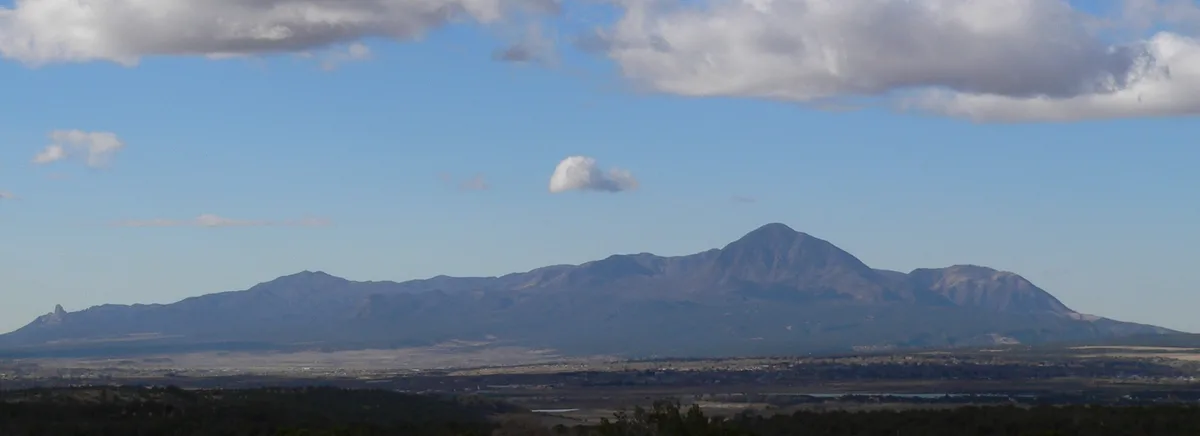 Sleeping Ute Mountain stretches across the horizon in a wide desert panorama, its long, sleeping-profile ridge rendered in soft blue-gray light. A few clouds float above the peak, with a dark foreground and pale valley floor emphasizing the mountain’s iconic silhouette.