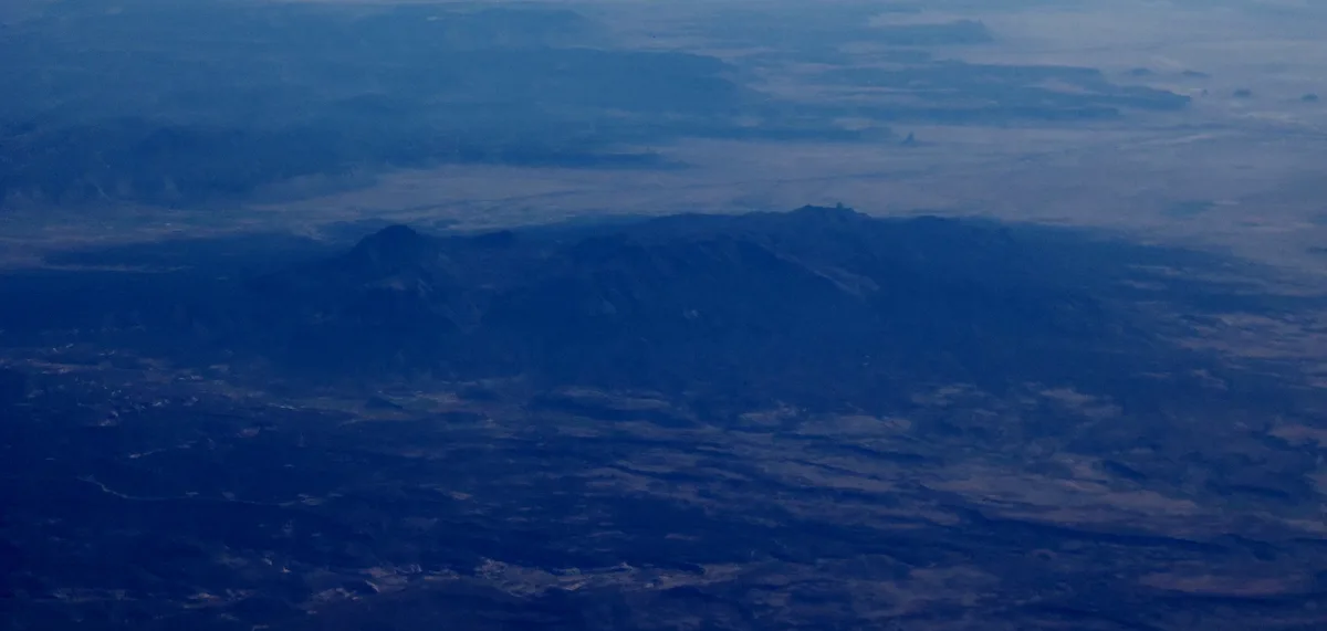 Sleeping Ute Mountain appears as a long, dark blue silhouette stretching across the center of the frame beneath a hazy sky. The wide landscape view shows the mountain rising from a muted desert plain, with soft evening light flattening the distant ridges and clouds.