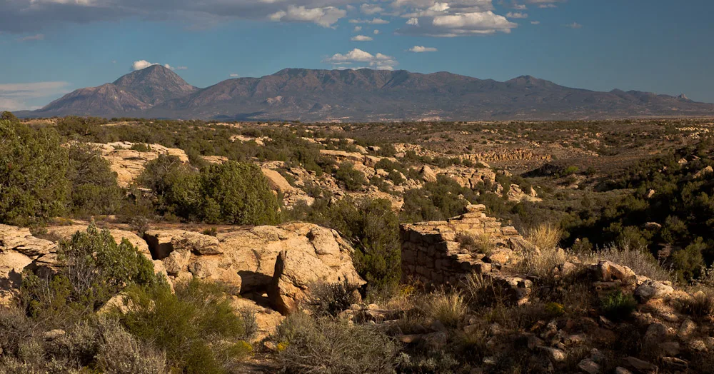 A wide desert view looks down a rocky canyon dotted with scrub and low trees toward Sleeping Ute Mountain, its long, dark silhouette stretching across the horizon under a few small clouds. Warm golden light picks out the tan sandstone ledges and layered terrain in the foreground.