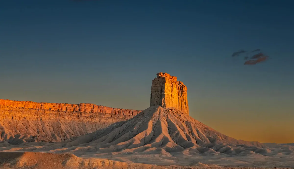 Sleeping Ute Mountain rises in a dramatic desert landscape, its flat-topped silhouette glowing orange in golden hour light against a deep blue sky. Eroded gray hills spread across the foreground, with a long sunlit cliff line stretching behind the mountain.