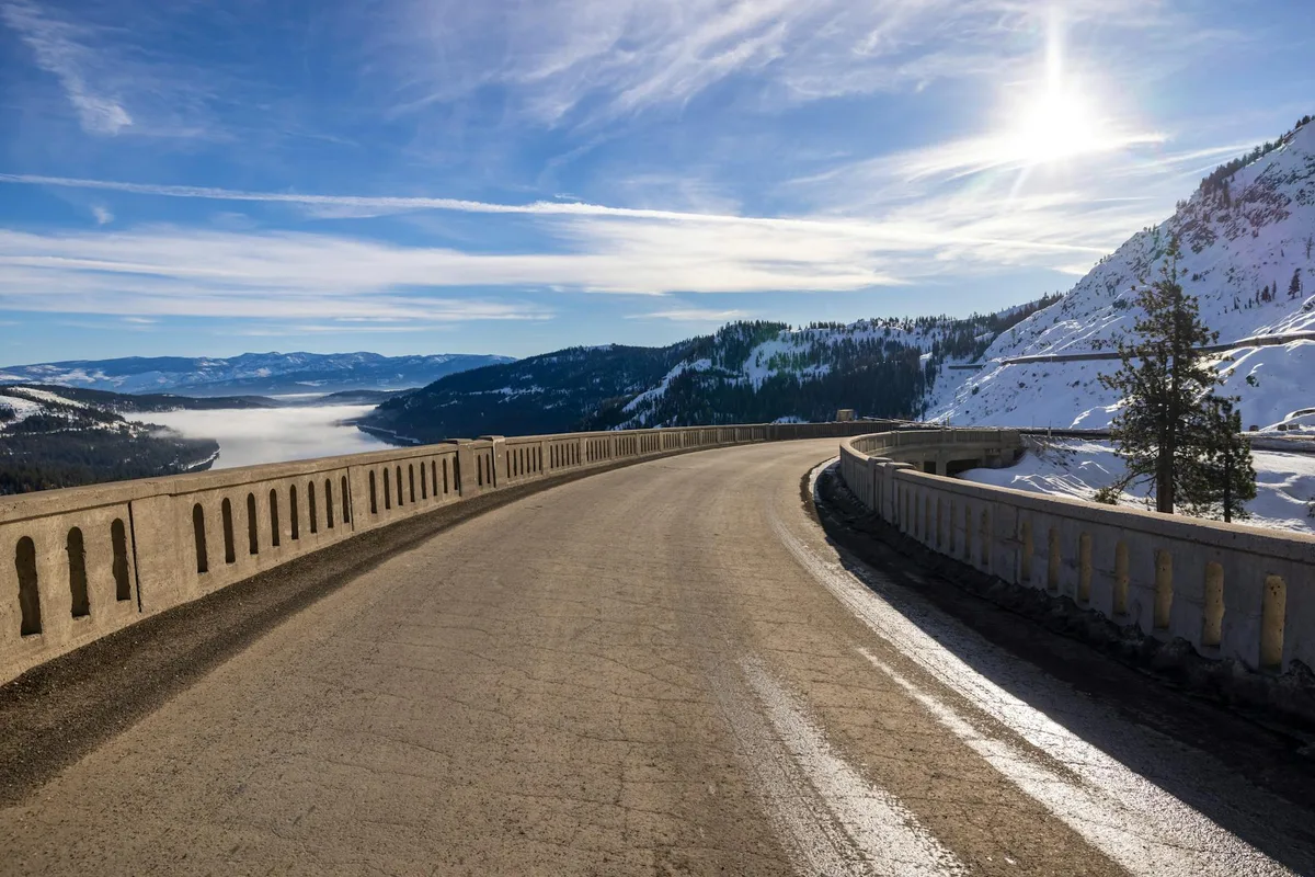 A curved mountain roadway with concrete guardrails leads toward a snowy overlook, with the bright afternoon sun glaring through thin clouds. Beyond the road, forested ridges and a low ribbon of mist fill the valley under a wide blue sky.