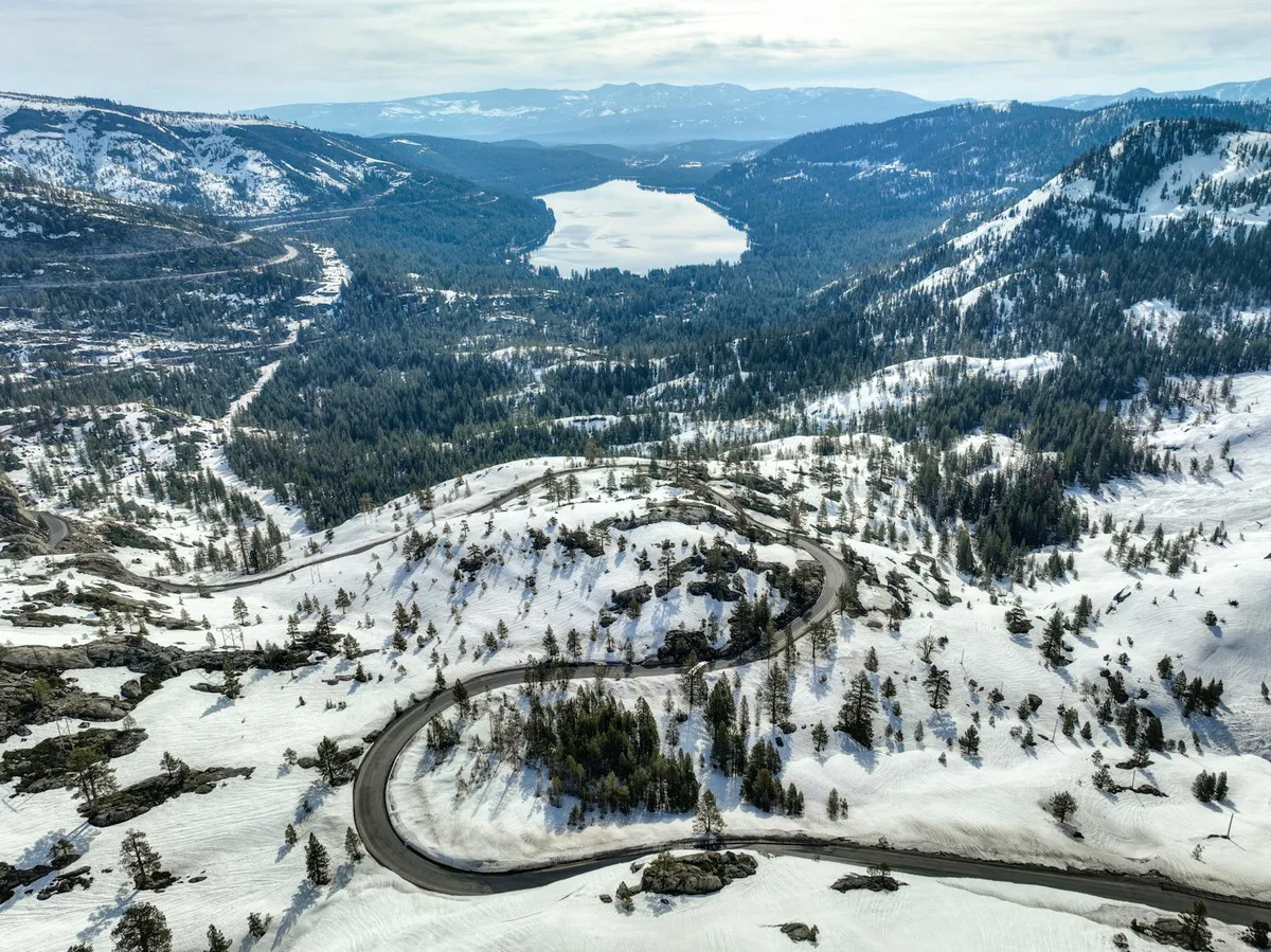 Snow-covered mountain slopes and dark evergreen forest fill the valley, with a winding scenic road curving through the foreground toward a bright lake in the distance. The high overlook view shows the broad, layered terrain under soft afternoon light.