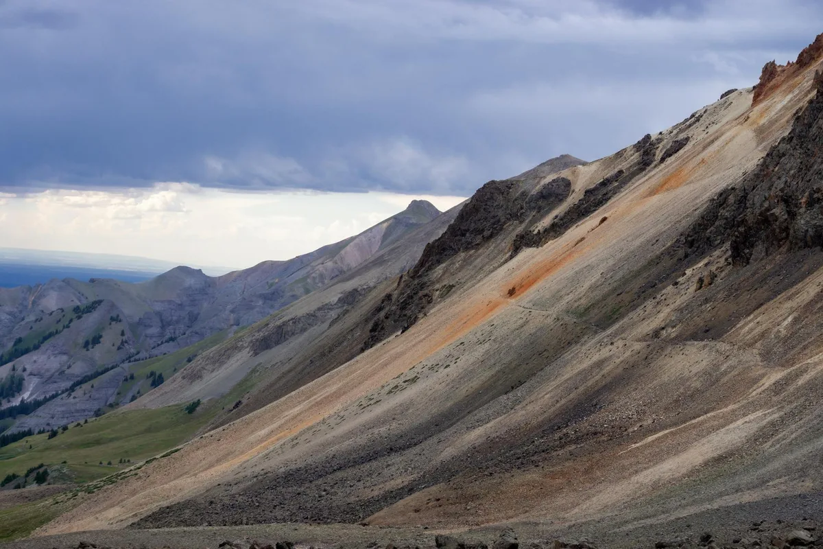 A steep, striped earthflow slopes across the mountainside, with pale gray and tan sediment cut by a vivid orange streak and dark rock outcrops. Green valley meadows and rugged peaks recede into the distance under a heavy, cloud-filled afternoon sky.