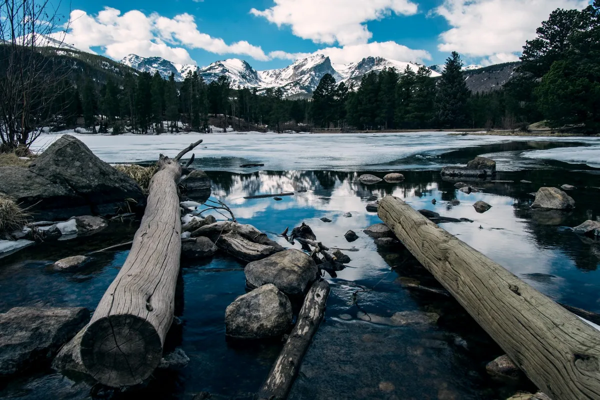 Driftwood and scattered rocks line the icy edge of Sprague Lake, where a dark patch of open water reflects the snowy peaks and trees beyond. Snow-covered mountains rise under a bright blue sky with broken clouds, giving the scene a cold, early-season feel.