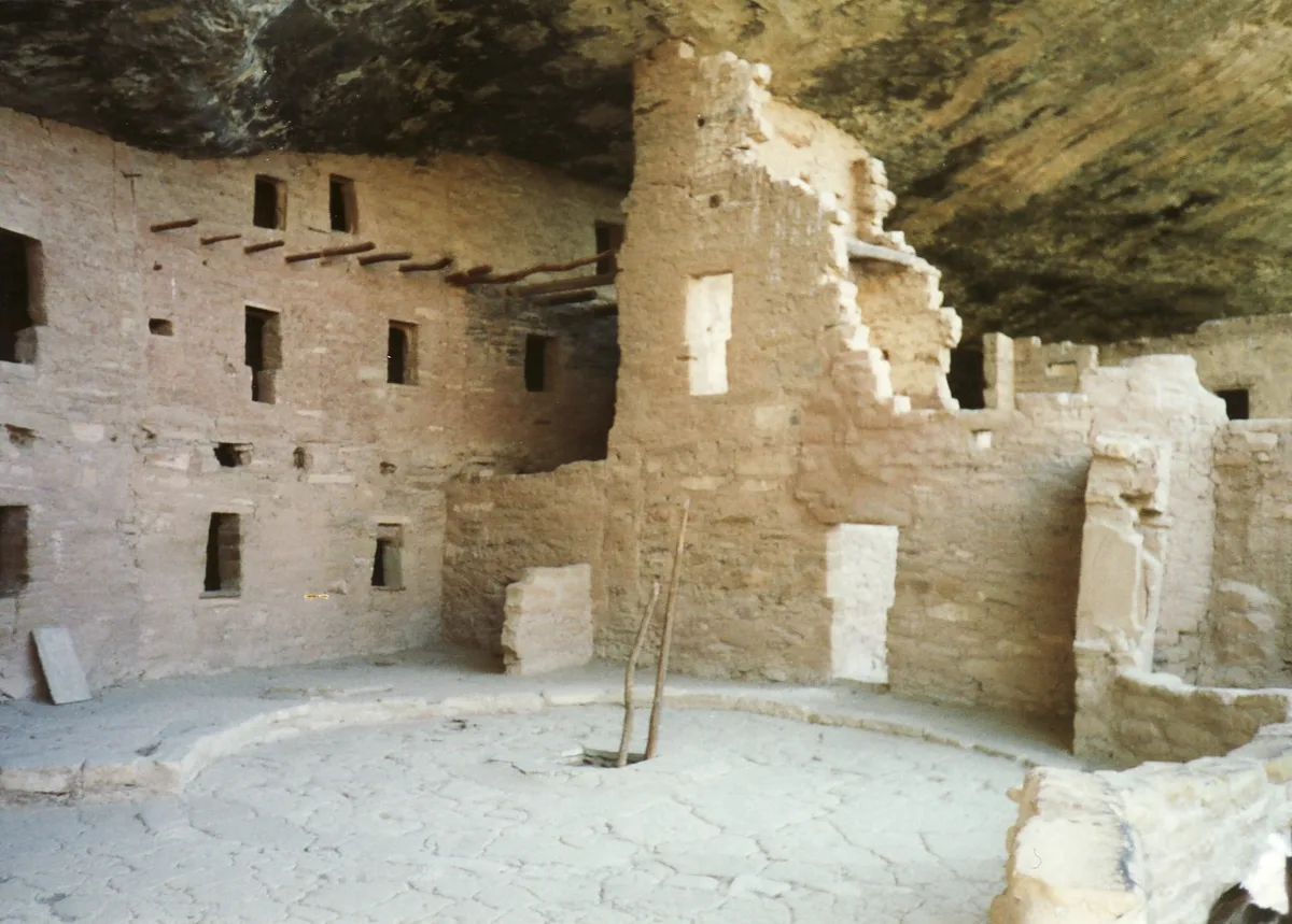 Inside a deep sandstone alcove, the cliff dwelling’s pale adobe-and-stone walls rise around a cracked, circular kiva floor, with dark window openings and exposed wooden roof beams. The overhanging rock ceiling presses low above the ruins, casting the sheltered interior in soft, muted light.