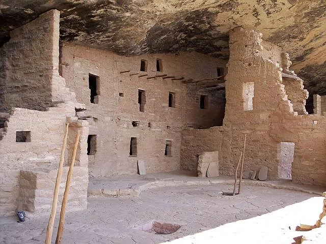 Ancient sandstone rooms and kivas sit beneath a deep rock overhang, with stacked masonry walls, square doorways, and weathered stone surfaces filling the cliff alcove. The morning light brightens the dusty floor and the right side of the ruins, while the dark ceiling of the alcove frames the dwelling above.
