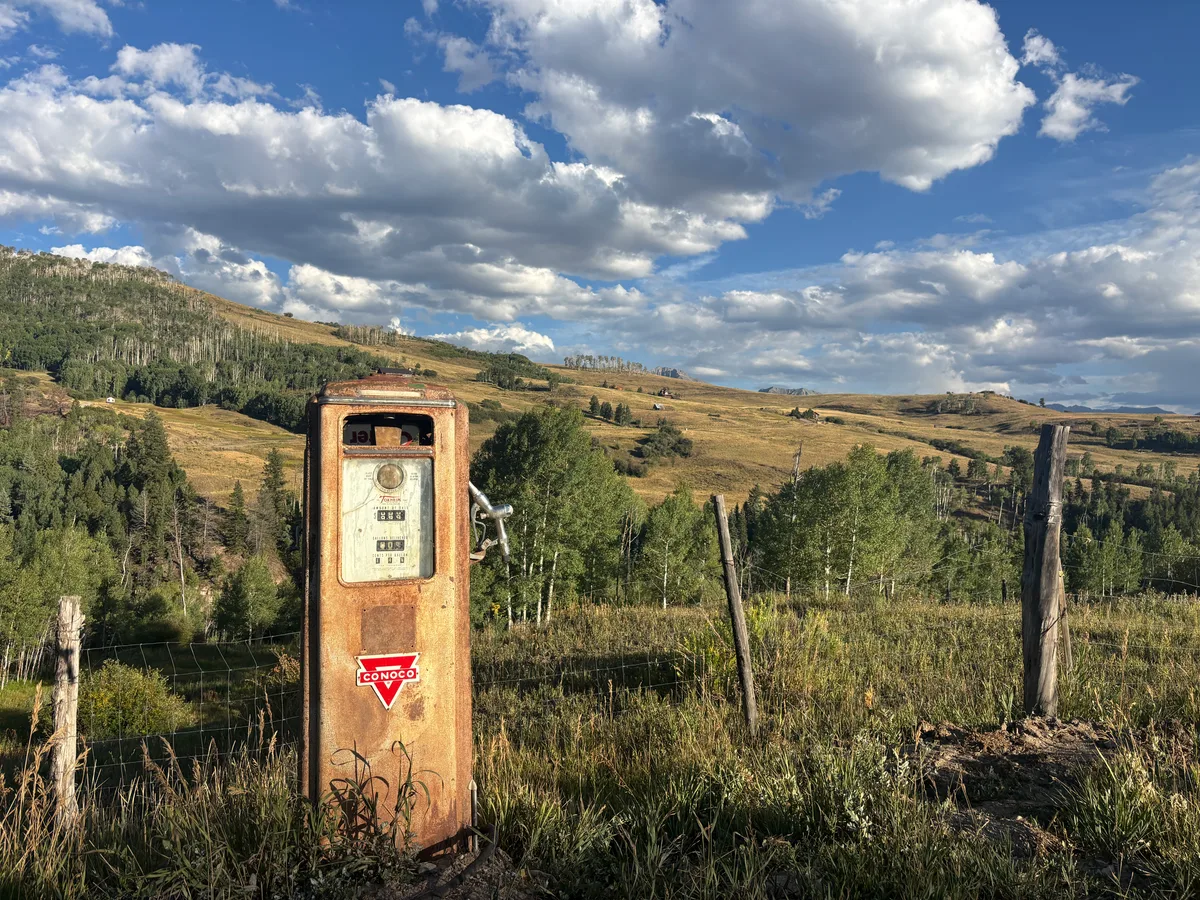 A rusted vintage Conoco gas pump stands in the foreground of a grassy hillside, with weathered fence posts and aspen-covered slopes stretching into the distance. Puffy cumulus clouds drift across a bright blue sky above the open mountain valley.