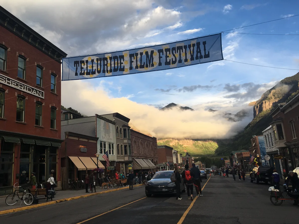 Colorado Avenue is lined with historic brick storefronts and pedestrians under a banner reading “Telluride Film Festival,” with low clouds glowing over the mountain peaks at the end of the street. The scene captures Telluride’s Victorian downtown framed by the steep box canyon walls in soft golden light.