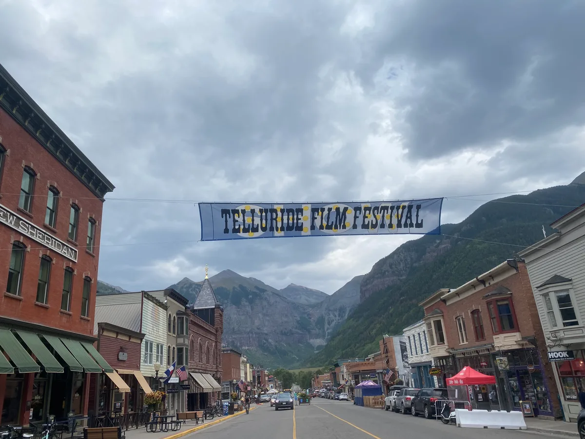 Historic Colorado Avenue in Telluride stretches toward steep, forested mountain walls under a cloudy sky, with the Telluride Film Festival banner hanging across the street. Victorian brick storefronts, awnings, parked cars, and small festival tents line the road, framing the narrow box canyon beyond.